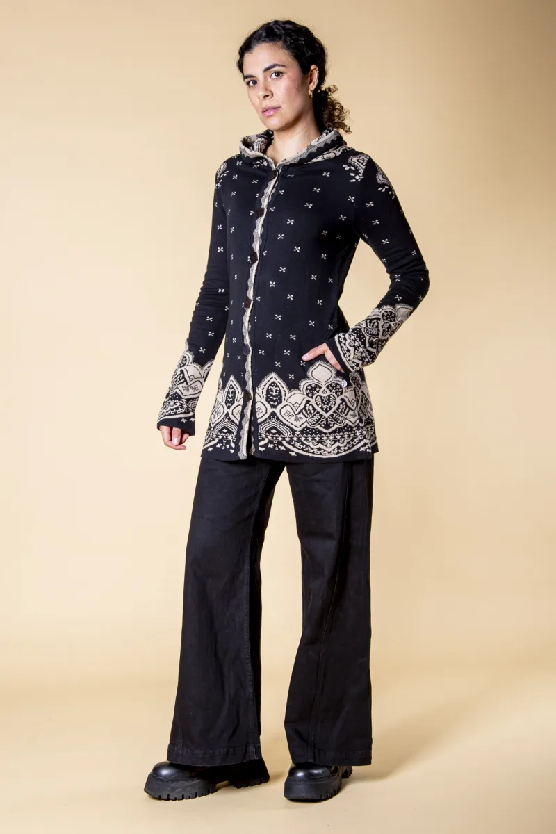 Model in einem schwarzen Mantel aus Strick-Jacquard mit kleinem cremefarbenem Muster, das an Ärmeln und Saum in ein großes Ornament übergeht, Modell Mavius