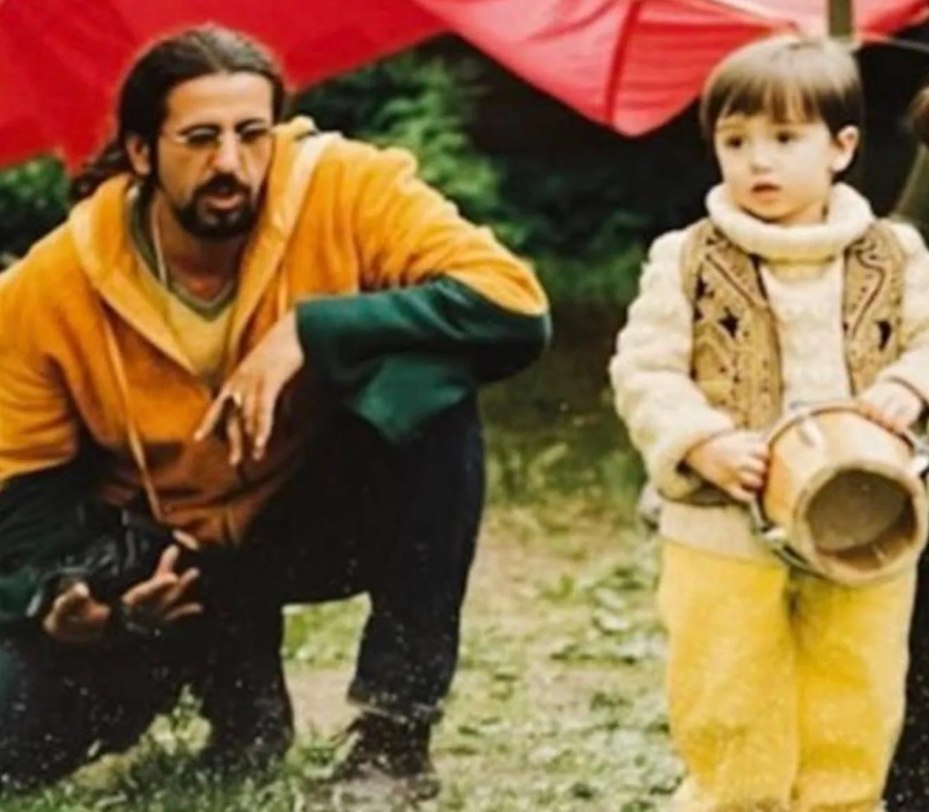 Hamid in gelb-grüner Kapuzenjacke hockt im Gras neben einem seiner Kinder, das eine kleine Trommel hält