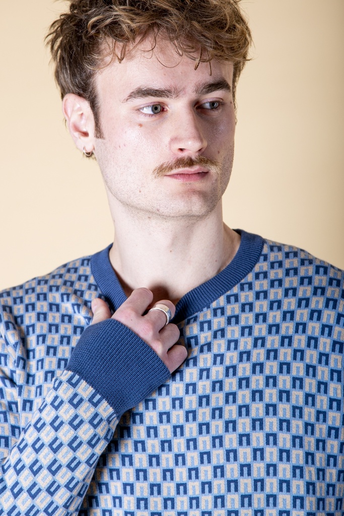 Nahaufnahme von Ausschnitt und Ärmel: der hellblaue Pullover aus Strick-Jacquard hat eine blaue Rippblende und ein feines Rechteckmuster.