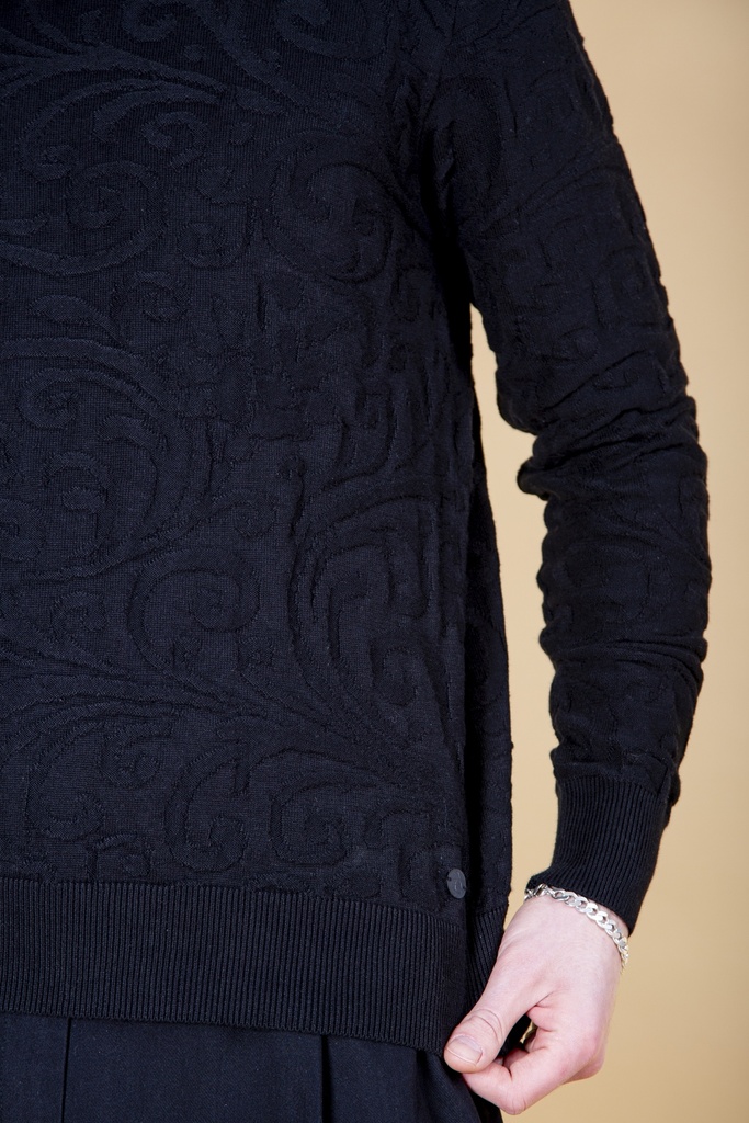 Nahaufnahme der Seite: der schwarze Pullover aus Strick-Jacquard zeigt Rankenmuster, langen Seitenschlitz mit kleinem Knopf und Rippbund.