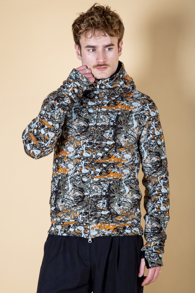 Vorderansicht bis zur Hüfte: Jacke aus Strick-Jacquard mit orange-grau-schwarzem Fleckenmuster, Reißverschluss, hohem Kragen und Ärmelstulpen.