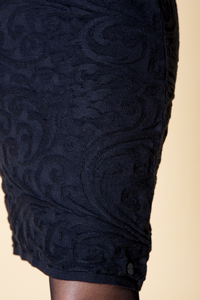 Nahaufnahme der Hüfte: am schwarzen Kleid aus Strick-Jacquard heben sich reliefartige Ornamente ab, eine kleine runde Markierung sitzt am Saum.