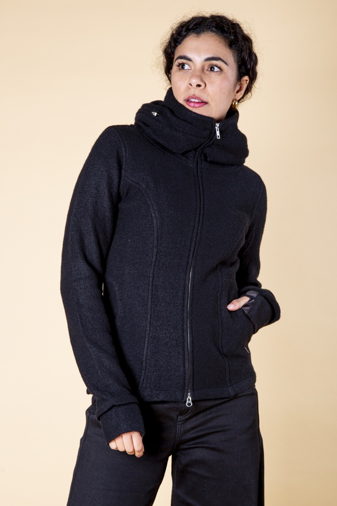 Oberkörper halbnah: schwarze Wolljacke mit Reißverschluss und breitem, schalartig gelegtem Kragen, eine Hand in der seitlichen Tasche.