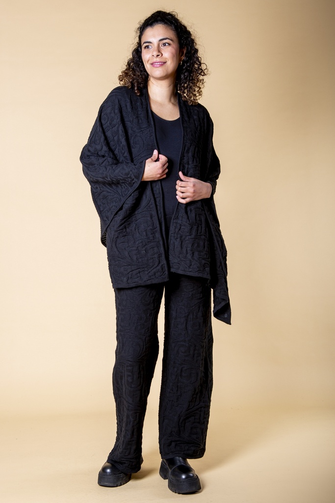 Vorderansicht in ganzer Länge: der offene schwarze Poncho fällt über die Hüfte, die gerippte Strickstruktur zeichnet sich auf den Hosen ab.