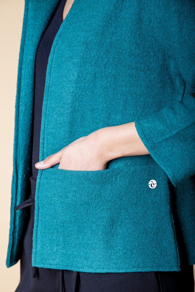 Nahaufnahme der Seite: eine Hand liegt in der petrolblauen Eingrifftasche mit Ziernaht und kleinem Knopf neben der offenen Vorderkante.