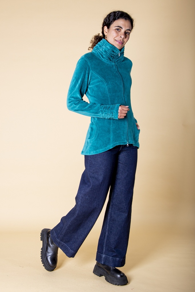 Seitliche Ganzkörperansicht: die türkisfarbene Samtjacke folgt der Taille, der hohe Kragen und die gerafften Stulpen zeichnen sich ab.