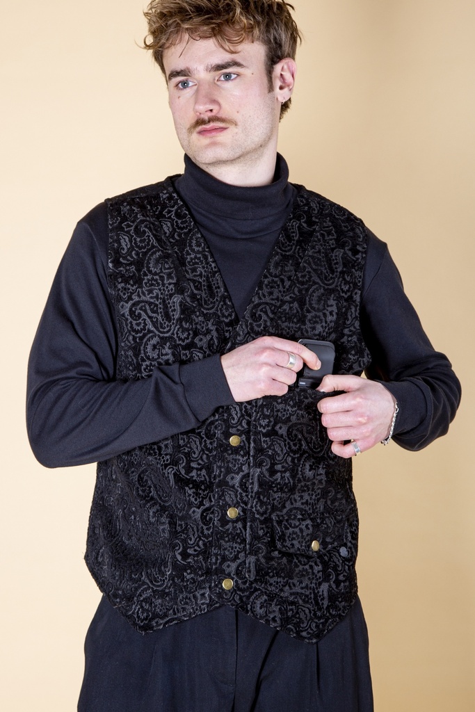 Vorderansicht bis zur Hüfte: die schwarze Paisley-Brokatweste ist geöffnet, die Druckknopfleiste und die Eingrifftaschen liegen über einem dunklen Rollkragen.