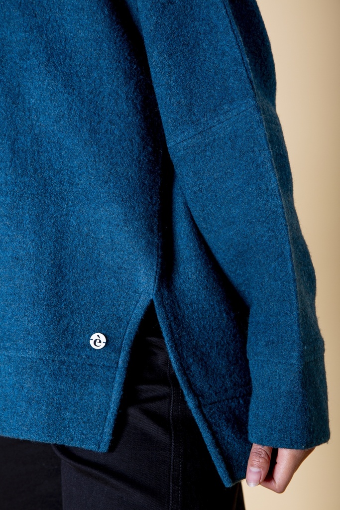 Nahaufnahme der Seite: ein langer Seitenschlitz öffnet den petrolblauen Pullover über der Hüfte, die Kante fällt unter dem breiten Ärmel gerade herab.