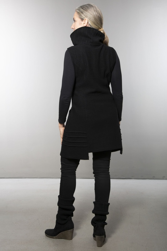 Ganzkörperansicht von vorn: lange schwarze Weste aus Wollstrick mit Reißverschluss und hohem Stehkragen, dazu ein dunkles Langarmshirt.