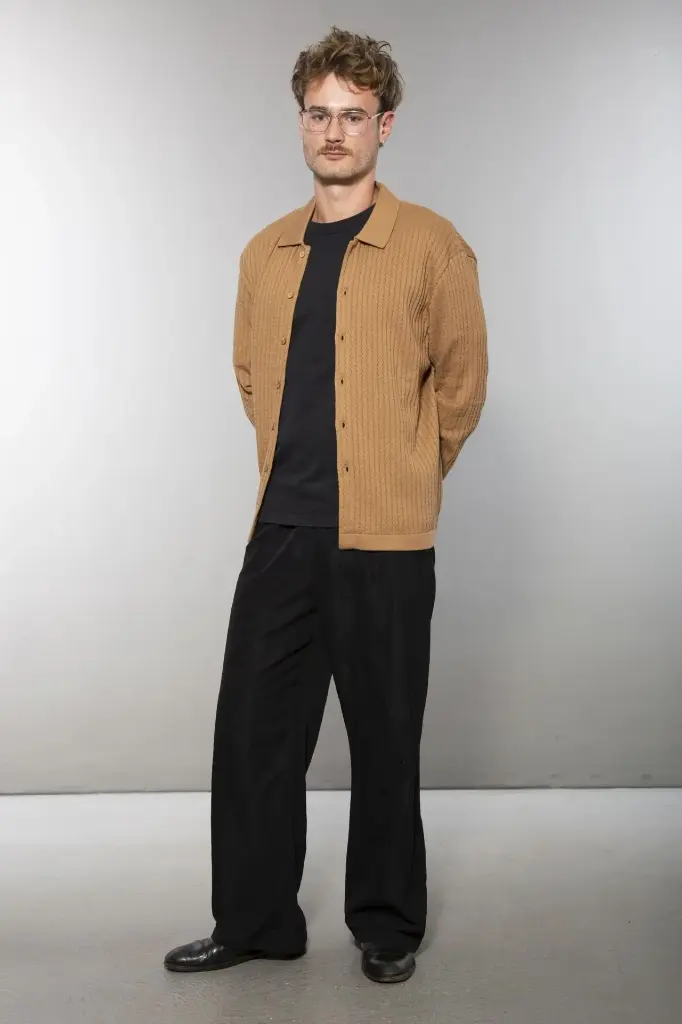 Ganzkörperansicht von vorn: schwarze Strickjacke mit Hemdkragen und Knopfleiste, senkrechte Rippenstruktur, dazu eine dunkle weite Hose.